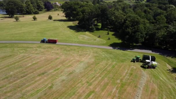 イギリスの田舎の公園でトレーラーを牽引するトラクターの空中ドローンショットと サイレージのための草の切り傷を収集するフォージ収穫機をサポートします — ストック動画