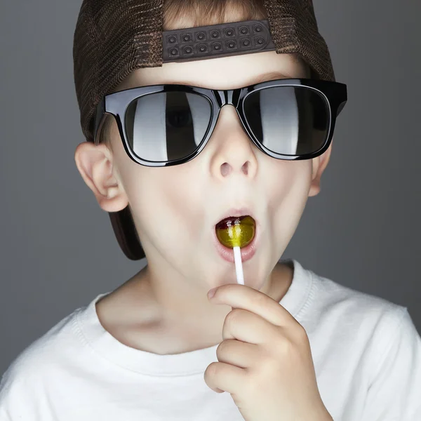 Niño divertido comiendo una piruleta. Niño de moda en gafas de sol —  Fotos de Stock