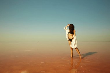 Güzel bir kız pembe bir tuz gölünün üzerinde suyun içinde duruyor. Bikinili genç kadın rahatlıyor.