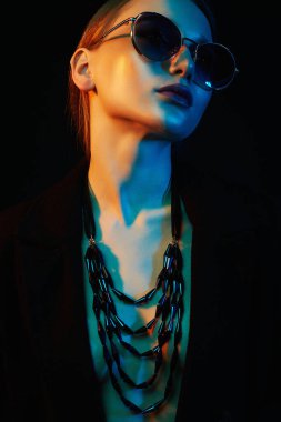 Renkli parlak ışıklar içinde güzel bir genç kadın. Güneş gözlüklü ve mücevherli güzel bir kızın moda portresi.