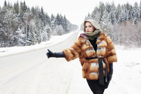 Hermosa chica en un abrigo de piel esperando el coche en una carretera de invierno en la mujer forest.young en abrigo de piel de moda, guantes de cuero y bufanda —  Fotos de Stock