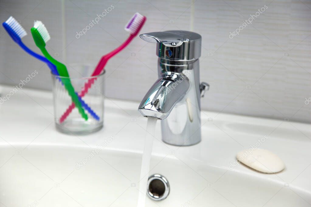 Toothbrushes, soap, and water tap faucet with running clean water in a white bathroom sink.