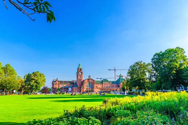 Wiesbaden Hauptbahnhof merkez tren istasyonu Neo-barok tarzı bina ve Reisinger-Anlagen parkı tarihi şehir merkezinde yeşil ağaçlar ve çimenler, mavi gökyüzü arka planı, Hesse eyaleti, Almanya