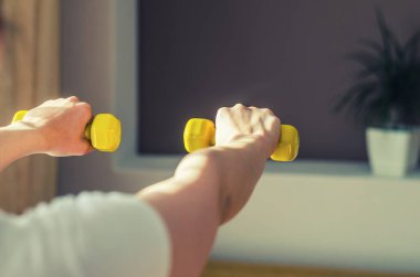 Young woman arms raising yellow dumbbells during workout sports at home, flower pot with green plant in wall niche in living room background, close-up view from back, sun light from side