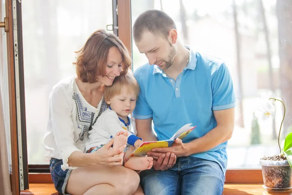 Familia feliz sentada en un alféizar de la ventana y leyendo un libro . — Foto de Stock