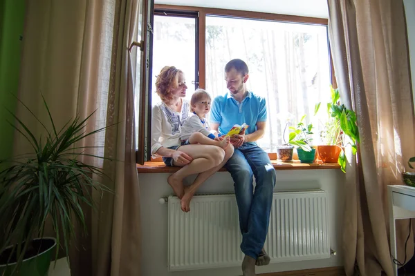 Familia feliz sentada en un alféizar de la ventana y leyendo un libro . — Foto de Stock