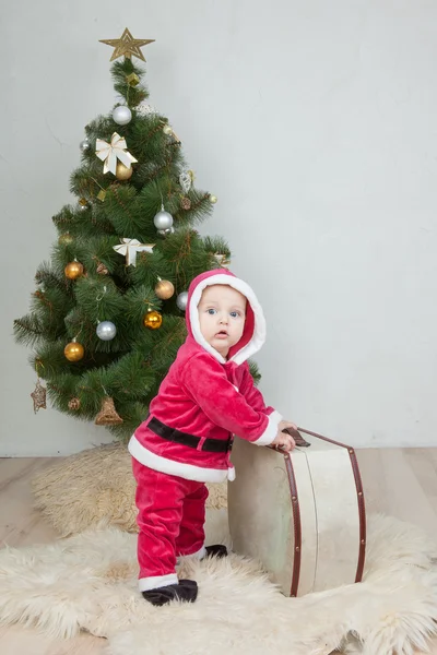 Menino pequeno no terno de Santa joga perto da árvore de ano novo no estúdio branco — Fotografia de Stock