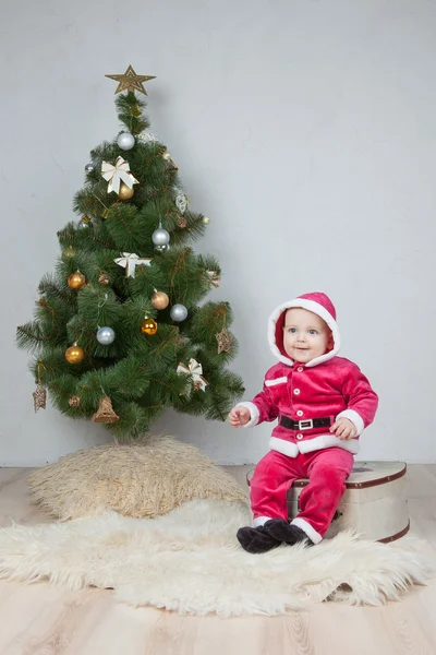 Menino pequeno no terno de Santa joga perto da árvore de ano novo no estúdio branco — Fotografia de Stock