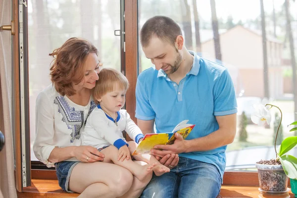 Familia feliz sentada en un alféizar de la ventana y leyendo un libro . — Foto de Stock