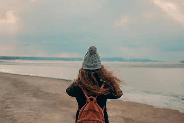 若い巻き毛の女の子の旅行者は ニット帽とバックパックでビーチに立っています — ストック写真