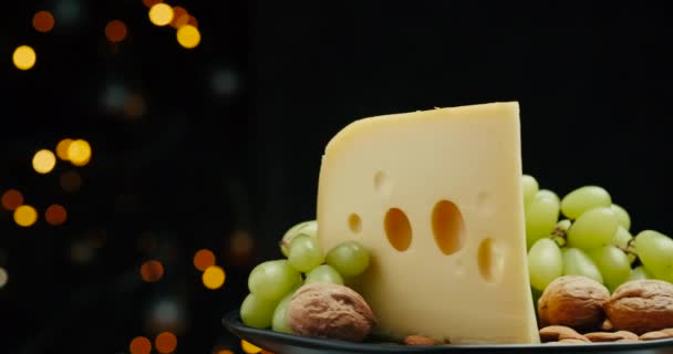 Κοντινό μέρος του Medium hard cheese head edam gouda παρμεζάνα σε ξύλινη σανίδα, με ξηρούς καρπούς και σταφύλια ροτάζ στο πιάτο. — Αρχείο Βίντεο