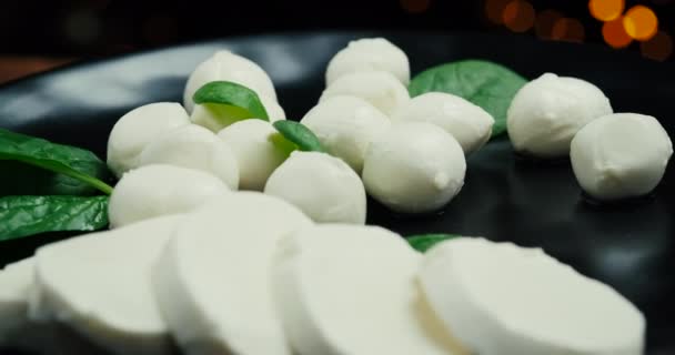 Witte kleine mozzarella kaasballen, spinazie bladeren op zwarte plaat. Roterend schot. — Stockvideo