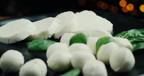 Witte kleine mozzarella kaasballen, spinazie bladeren op zwarte plaat. Roterend schot. — Stockvideo