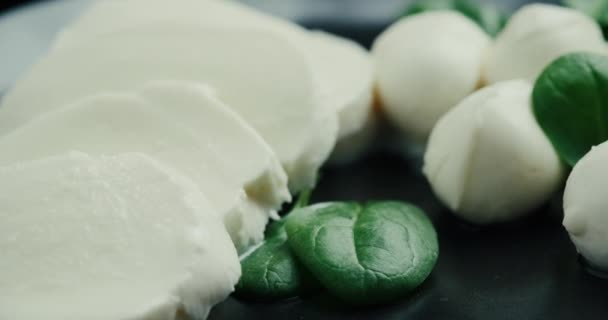 Vita små mozzarella ostbollar, spenatblad på svart tallrik. Rotationsskott. — Stockvideo