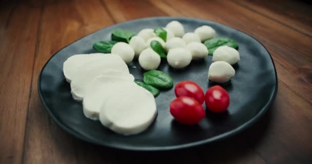 白い小さなモッツァレラチーズボール,ほうれん草の葉と黒プレート上のトマト.ドリーインショット. — ストック動画