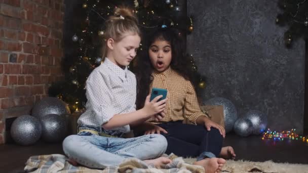 Két csinos fiatal afrikai és kaukázusi lányok ül közel egy újévi vagy karácsonyfa és kommunikálni videokapcsolat egy okostelefon. — Stock videók