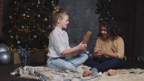 Adolescentes felices intercambiando regalos. Dos chicas africanas y caucásicas muy jóvenes sentadas cerca de un árbol de Año Nuevo o de Navidad y se regalan mutuamente. — Vídeos de Stock