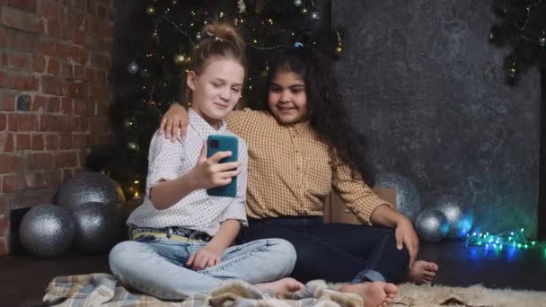 İki güzel Afrikalı ve Kafkasyalı kız yılbaşı ağacı ya da Noel ağacının yanında oturuyor ve akıllı telefondan video bağlantısı ile iletişim kuruyorlar.. — Stok video