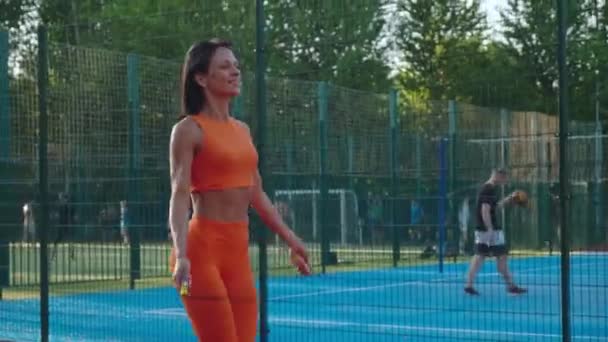 오렌지 운동복을 입은 젊은 여성이 줄을 줄넘기로 스포츠 경기장에서 점프를 한다. — 비디오