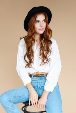 Siyah şapkalı, mavi kotlu, şeftali rengi arka planda oturan güzel, gülümseyen kızıl saçlı kız.