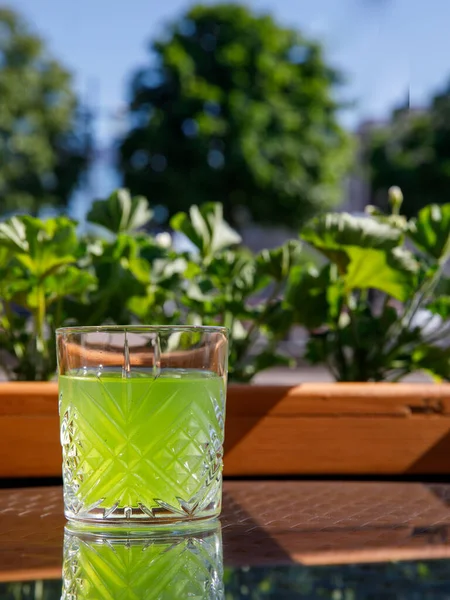 Glass with a bright green chilled drink on the table in a street outside cafe with a view of the sea in the sun day. Vertical, copy space, side view