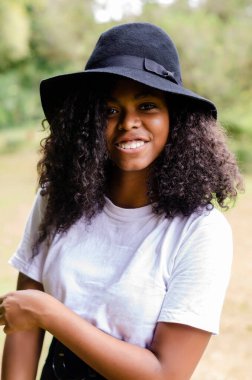 20-25 yaşlarında, genç, siyah bir kadın. Parkta kıvırcık saçlı, kameraya bakıyor.