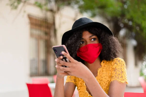 young black woman with curly hair, with red mask, yellow dress and black hat, checking her phone sitting in restaurant