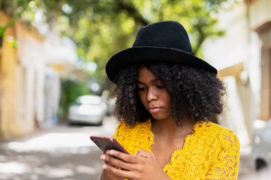 young black woman with curly hair, with red mask, yellow dress, using her smart phone