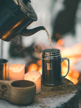 Çay çaydanlıktan kamp ateşi bardağına dökülür. Kışın ormanda bir ağaç kütüğünün üzerinde durur.