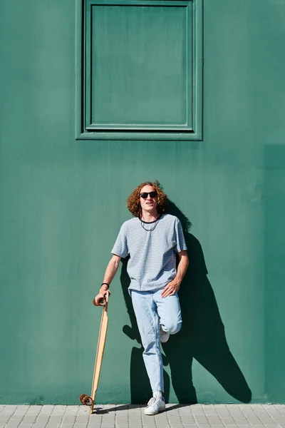 Ung man med lockigt hår med solglasögon och en longboard eller skridsko med en grön vägg bakom i en solig sommardag — Stockfoto