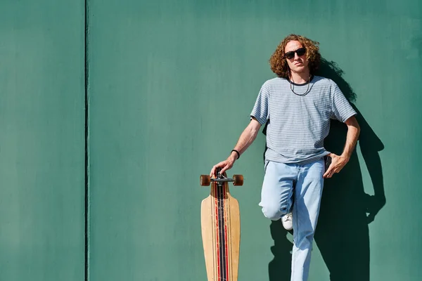 Un joven con el pelo largo rizado y gafas de sol con un longboard o patinar con una pared verde se comportan él —  Fotos de Stock