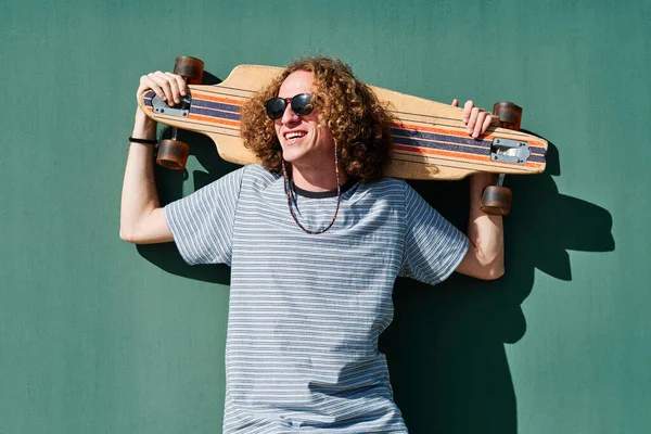 Un joven hipster mirando al sol con gafas de sol y un longboard o patín en los hombros —  Fotos de Stock