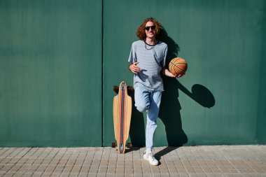 Kıvırcık saçlı, güneş gözlüklü, kaykaylı ve arkasında yeşil duvar olan bir basketbol topu olan genç bir adam.