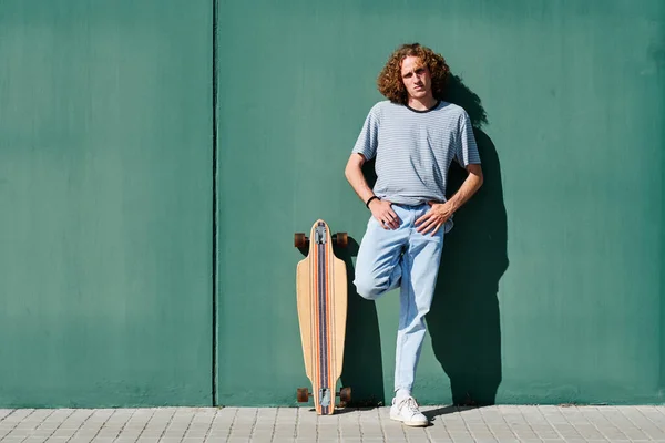 Un joven con el pelo rizado de pie en una pared verde en un día soleado con su patín o longboard —  Fotos de Stock