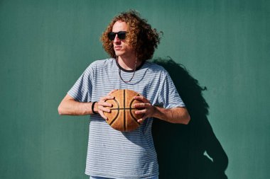 Basketbol topu ve güneş gözlüklü genç bir adam arkasında yeşil bir duvar olan güneşe bakıyor.