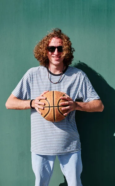 Un joven con el pelo rizado y una pelota de baloncesto mirando a la cámara con una pared verde detrás de él —  Fotos de Stock