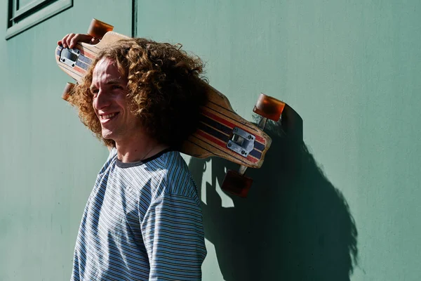 Un joven con el pelo rizado y gafas de sol con su patín en los hombros —  Fotos de Stock