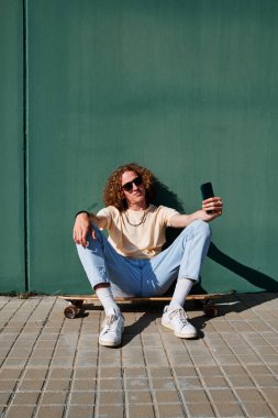 Akıllı telefonunu kullanan genç bir adam kaykayına ya da sörf tahtasına oturmuş ve arkasında bir duvar var.