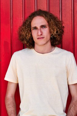 Kıvırcık saçlı, yeşil gözlü, kırmızı arka planda kameraya bakan ciddi bir gencin portresi.