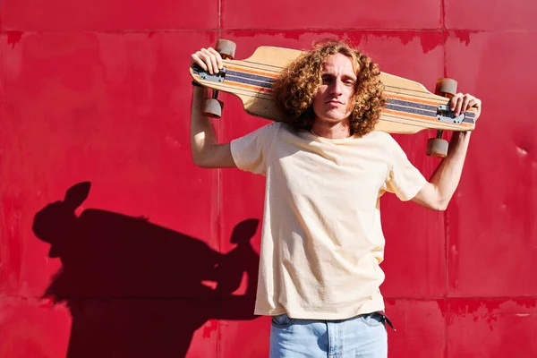 Joven con el pelo rizado mirando a la cámara con su monopatín en los hombros y un fondo rojo —  Fotos de Stock