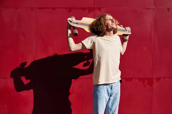 Joven y elegante hombre con el pelo rizado mirando al sol con su monopatín en los hombros y con un fondo rojo —  Fotos de Stock