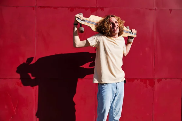 Joven con el pelo rizado mirando a la cámara con su monopatín en los hombros y un fondo rojo —  Fotos de Stock