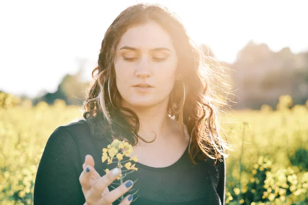 Jeune femme courbée avec une fleur de colza dans un champ de colza au printemps avec le soleil derrière elle — Photo