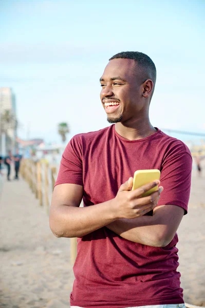 Jonge zwarte man aan het strand glimlachend en met behulp van zijn smartphone — Stockfoto