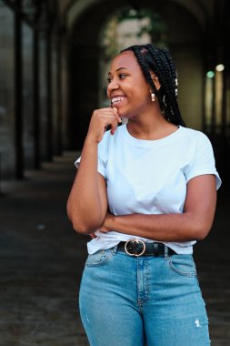 Saçları örgülü genç Latin bir kadın açık havada günlük giysiler içinde gülümsüyor.