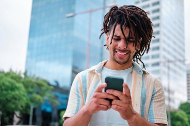 kıvırcık saçlı genç adam modern şehir binalarıyla çevrili akıllı telefon kullanıyor.