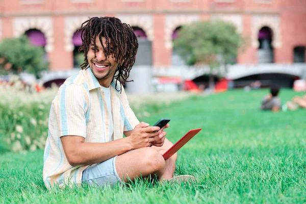 Joven estudiante sonriendo y utilizando el teléfono inteligente y ordenador portátil en el parque —  Fotos de Stock