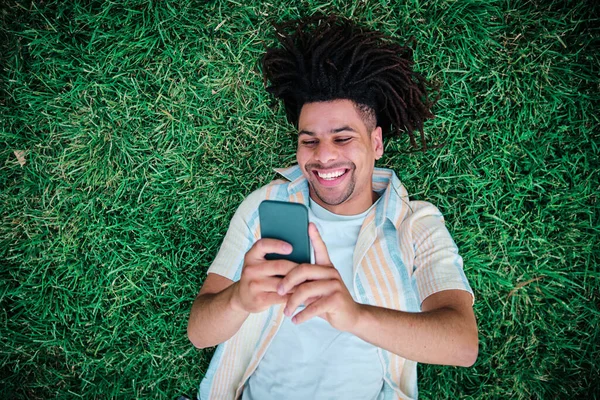 스마트폰을 사용하여 잔디에 누워 젊은 곱슬머리 라틴 남자 — 스톡 사진