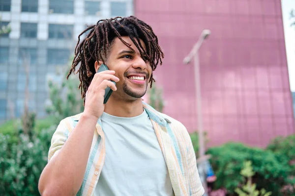 Joven latino con el pelo rizado con el horizonte de la ciudad en el fondo usando el teléfono inteligente y sonriendo —  Fotos de Stock