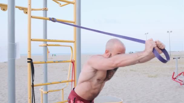 Atletische mannentraining met een elastiekje aan het strand. Calisthenisch trainingsconcept. — Stockvideo